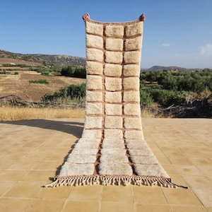 Op de afbeelding: Een rechthoekig, crèmekleurig tapijt met een rasterpatroon van verhoogde vierkanten en bruine randen. Het tapijt heeft een gefranjerde rand en wordt buiten gepresenteerd tegen een achtergrond van een veld en een blauwe lucht. Het tapijt is gemaakt van een zacht, getextureerd materiaal.