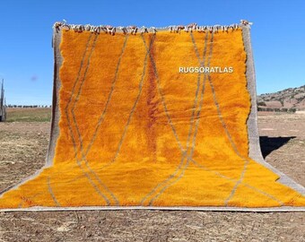 May include: A large, rectangular, orange rug with a geometric design of gray lines. The rug is displayed outdoors against a backdrop of a clear blue sky and a rural landscape. The text "RUGSORATLAS" is visible on the rug.