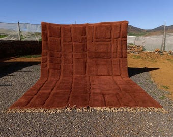 May include: A large, rectangular, rust-colored rug with a geometric pattern of raised squares and rectangles. The rug has a fringe along the bottom edge and is displayed outdoors on a gravel surface.