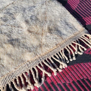 May include: Close-up of a beige and gray textured rug with a cream-colored fringe. The rug has a shaggy appearance and is placed on top of a patterned magenta and black surface. The rug's texture is visible.