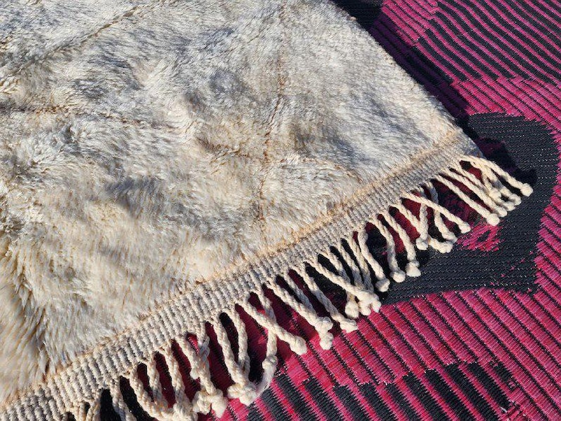 May include: Close-up of a beige and gray textured rug with a cream-colored fringe. The rug has a shaggy appearance and is placed on top of a patterned magenta and black surface. The rug's texture is visible.