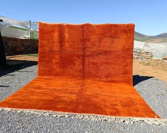 May include: A large, rectangular orange rug with a textured surface and a cream-colored fringe. The rug is displayed outdoors on a gravel surface, with a glimpse of a tent and a mountain range in the background. The rug's dimensions are not visible.