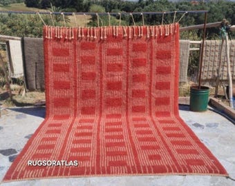 May include: A large, rectangular red rug with a geometric pattern of vertical stripes and rectangular blocks. The rug is displayed outdoors, with the text "RUGSORATLAS" visible at the bottom. The rug has a fringe along the top edge.