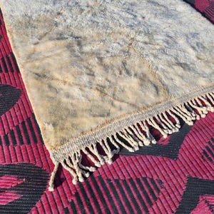 May include: A rectangular rug with a cream-colored, textured surface and a fringe border. The rug is placed on a red and black patterned surface, creating a contrast in textures and colors.