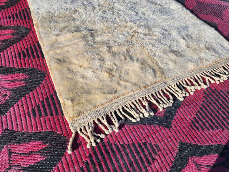 May include: A rectangular rug with a cream-colored, textured surface and a fringe border. The rug is placed on a red and black patterned surface, creating a contrast in textures and colors.