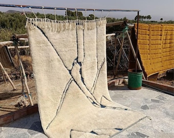 May include: A cream-colored, rectangular rug with a geometric design of dark blue lines. The rug is hanging outside, with a fringe along the top edge. Another rug is visible in the background, along with a green bucket.