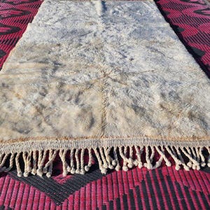 May include: A rectangular, off-white rug with a shaggy texture and a fringe border. The rug has a light brown center and is placed on a red and black patterned surface. The rug's dimensions are not visible.