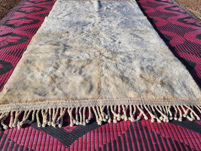 May include: A rectangular, off-white rug with a shaggy texture and a fringe border. The rug has a light brown center and is placed on a red and black patterned surface. The rug's dimensions are not visible.