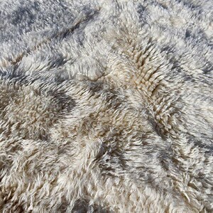 May include: Close-up of a textured, off-white rug with a shaggy, layered appearance. The rug has a mix of light and dark tones, creating a soft, inviting look. The fibers are visible, adding depth and dimension.