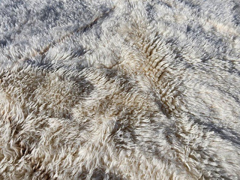 May include: Close-up of a textured, off-white rug with a shaggy, layered appearance. The rug has a mix of light and dark tones, creating a soft, inviting look. The fibers are visible, adding depth and dimension.