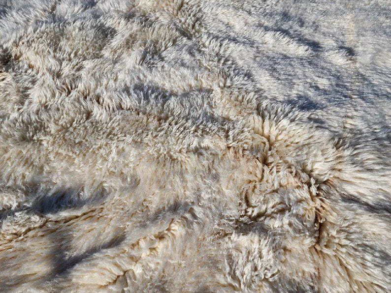 May include: Close-up of a textured, off-white rug with long, shaggy fibers. The rug has areas of light and shadow, creating a soft, natural look. The fibers are thick and appear to be made of wool.