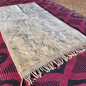 May include: A rectangular, off-white rug with a shaggy texture and a fringe along one edge. The rug has a faded appearance with brown and beige tones. The rug is placed on a red and black patterned surface.