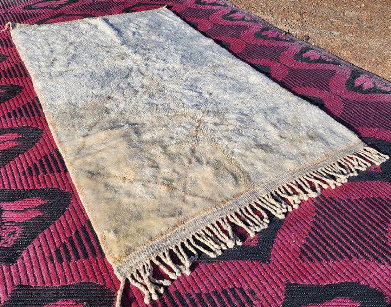 May include: A rectangular, off-white rug with a shaggy texture and a fringe along one edge. The rug has a faded appearance with brown and beige tones. The rug is placed on a red and black patterned surface.