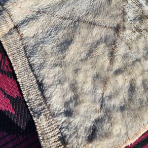 May include: Close-up of a textured, beige and gray faux fur blanket. The blanket has a soft, plush appearance with darker gray patches. A red and black patterned mat is visible in the corner.
