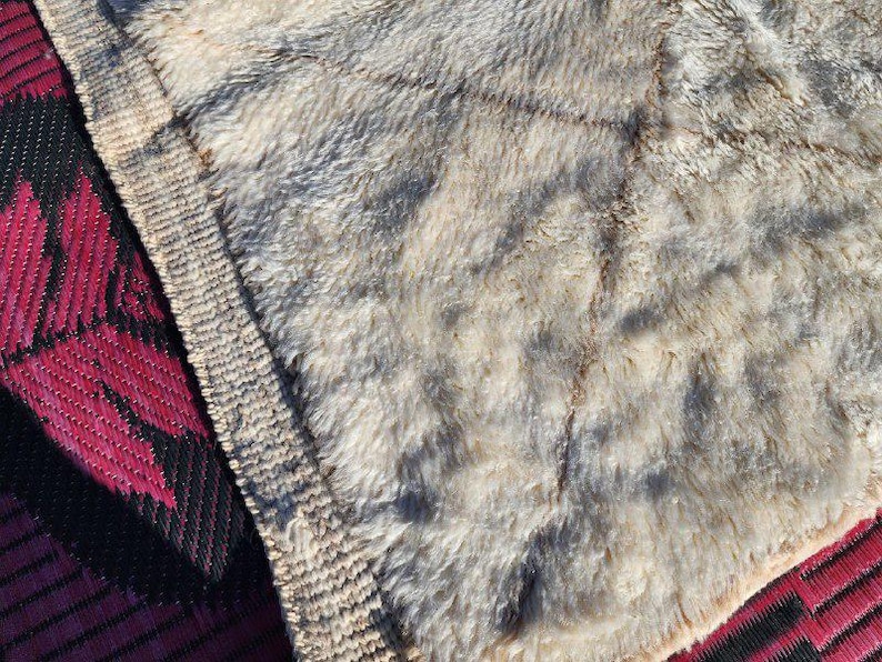 May include: Close-up of a textured, beige and gray faux fur blanket. The blanket has a soft, plush appearance with darker gray patches. A red and black patterned mat is visible in the corner.