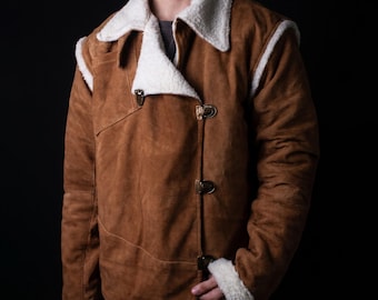 May include: A tan suede jacket featuring a white faux shearling collar and cuffs. The jacket has an asymmetrical front closure secured with three metallic toggle clasps. The design includes visible seam detailing across the chest and shoulders.
