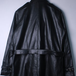 May include: A black leather trench coat hanging on a metal hook. The coat has a belt at the waist, shoulder epaulettes, and buttoned cuffs. The leather has a slightly textured appearance.