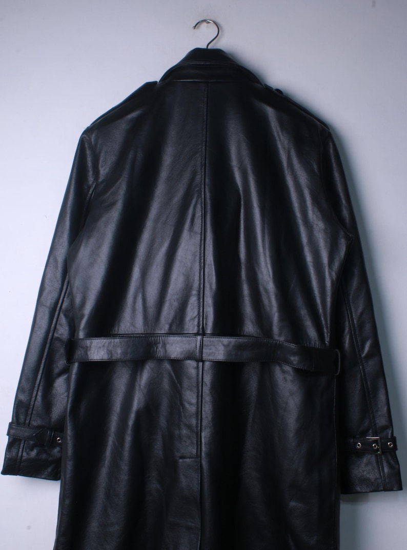 May include: A black leather trench coat hanging on a metal hook. The coat has a belt at the waist, shoulder epaulettes, and buttoned cuffs. The leather has a slightly textured appearance.