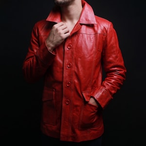 May include: A vibrant red leather jacket featuring a structured collar, long sleeves, and a front button closure. The garment includes two prominent rectangular patch pockets on the lower front section and visible vertical seam detailing throughout.