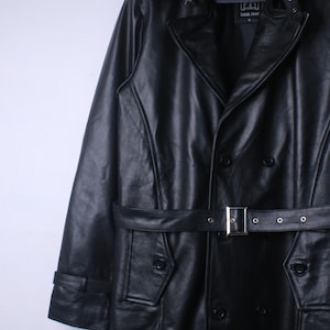 May include: Black leather jacket in a trench coat style, with a double-breasted front and a belt. The jacket has a notched collar, button details, and a tailored fit. The belt is fastened with a silver buckle.