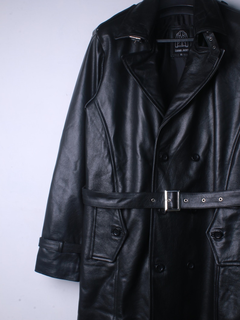 May include: Black leather jacket in a trench coat style, with a double-breasted front and a belt. The jacket has a notched collar, button details, and a tailored fit. The belt is fastened with a silver buckle.