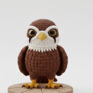 Puede incluir: Una figura de pájaro de ganchillo marrón hecha a mano con detalles blancos y negros, y patas y pico amarillos. El pájaro está posado sobre una pequeña base de madera redonda. El pájaro tiene ojos grandes y expresivos.