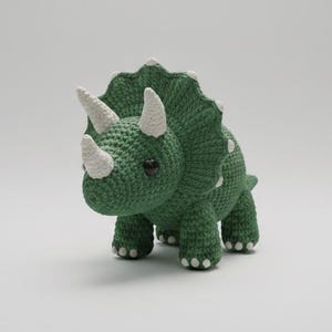 Könnte beinhalten: Ein handgefertigtes grünes Triceratops-Dinosaurier-Amigurumi. Der Häkeldinosaurier hat drei weiße Hörner, weiße Akzente auf seinem Kragen und kleine schwarze Augen. Der Dinosaurier steht auf vier Beinen.