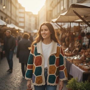 Puede incluir: Una mujer con una chaqueta de ganchillo de patrones geométricos coloridos, con cuello y ribetes crema, sobre una camiseta blanca. La chaqueta presenta parches hexagonales en tonos turquesa, naranja, marrón y morado. La escena tiene lugar en un mercado al aire libre.