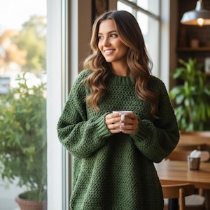 May include: A woman wearing a green knit sweater holds a white mug. The sweater has a textured pattern and long sleeves. She is smiling and looking out a window, with a blurred background of a cafe setting.