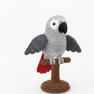 Peut inclure: Un perroquet crocheté avec un corps gris, des ailes gris foncé et une queue rouge. L'oiseau a un bec noir et des yeux jaunes. Il est perché sur un support crocheté marron avec une base ronde.
