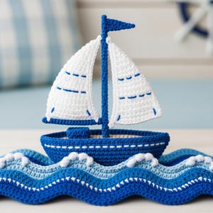 Puede incluir: Un velero de ganchillo hecho a mano en tonos azules y blancos. El barco presenta velas blancas con costuras azules, un mástil azul y un casco azul. Se asienta sobre un diseño de olas de ganchillo, creando un tema náutico. Una pequeña bandera azul está en la parte superior del mástil.