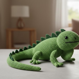 Puede incluir: Un juguete de iguana verde hecho a mano con ganchillo, con ojos negros y una boca sonriente. La iguana tiene una cola larga y una fila de púas triangulares en la espalda. El juguete está hecho de hilo suave.