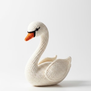 May include: A handmade crochet swan in off-white yarn. The swan has a long, curved neck, a black beak outline, and an orange beak. The wings are slightly spread, and the swan is sitting. A charming, handcrafted decorative item.