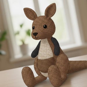 May include: A handmade crochet kangaroo plush toy in shades of brown, with a white belly and black arms. The kangaroo has large, expressive eyes and is sitting on a light-colored surface. The toy is detailed with a textured crochet stitch.