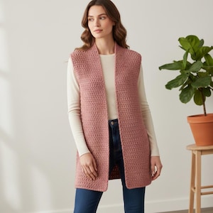 May include: A dusty rose-colored, open-front crochet vest. The vest is long, reaching to the mid-thigh, and has a simple, textured stitch pattern. The model is wearing the vest over a long-sleeved white shirt and blue jeans.