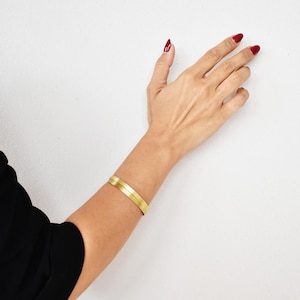 Op de afbeelding: Een goudkleurige armband met twee banden wordt om de arm van een persoon gedragen. De arm is tegen een witte achtergrond geplaatst. De nagels van de persoon zijn rood gelakt. De persoon draagt een zwarte mouw.