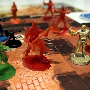 May include: A close-up of a game board with various miniature figures. The figures are made of plastic and are painted in different colours, including red, green, gold, and blue. The game board is made of cardboard and has a brown and orange colour scheme. The game board has the text "Sub Terra II" printed on it.