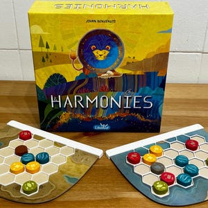 May include: A board game called "HARMONIES" is shown, with a yellow box featuring a lion illustration. Two game boards with hexagonal grids and colourful game pieces are also visible. The boards are brown and blue.