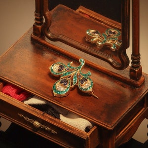 Op de afbeelding: Een kleine houten kaptafel met spiegel, lade en decoratieve details. Een pauwvormige broche met groene, blauwe en witte stenen ligt op het oppervlak. De lade is gedeeltelijk open en toont stof.