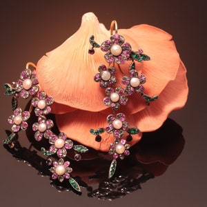 May include: Elegant floral drop earrings with gold hooks. Each earring features clusters of pink and clear gemstones surrounding a central pearl. Green accents add detail. The earrings are displayed against a peach-colored backdrop.