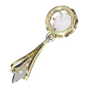 May include: A gold pendant with a round, clear gemstone set in a bezel setting. The pendant is attached to a delicate gold chain with a small, round gemstone at the bottom.