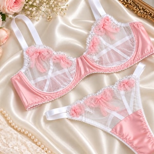 May include: A matching lingerie set consisting of a bra and thong in soft pink satin and white sheer mesh. The bra features white adjustable straps, white lace trim, and pink tulle rosettes. The thong includes matching pink rosettes and mesh.