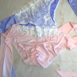 May include: Two pairs of lingerie. One pair is light pink satin with white lace trim and attached ribbons. The other pair is lavender with white lace trim. The lingerie is displayed on a light-colored surface.