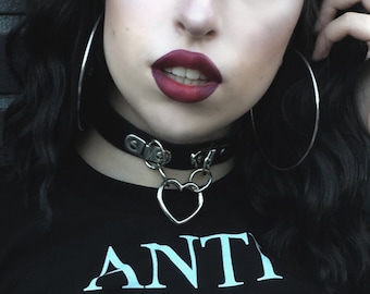 Può includere: Una maglietta nera con la scritta "ANTI DEMON DEMON CLUB" in bianco. L'immagine presenta anche un choker nero con un ciondolo a forma di cuore e grandi orecchini a cerchio argentati. La persona nell'immagine ha i capelli scuri e indossa trucco scuro.