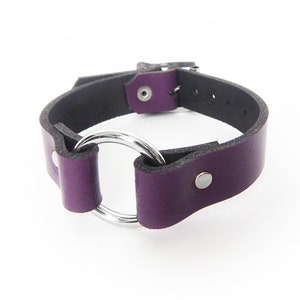Purple leather wristband with a silver coloured O ring in the center, and two silver coloured flat shaped rivets holding the purple leather and silver O ring in place.