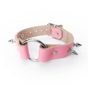 Könnte beinhalten: Rosa Lederhalsband mit silbernem Metallring und spitz zulaufenden Nieten. Das Halsband hat einen Schnallenverschluss und ist vor einem weißen Hintergrund platziert. Das Leder ist hellrosa.