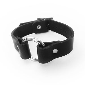 Black leather wristband with a silver coloured O ring in the center, and two silver coloured flat shaped rivets holding the black leather and silver O ring in place.