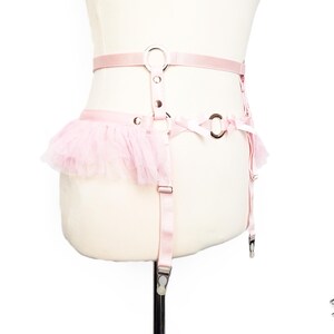 Adjustable pink vegan leather garter belt crafted for both functionality and style, catering to alternative fashion enthusiasts and BDSM communities alike. Featuring a central silver O ring, pink satin bow details, ruffled tulle accents at the side of the hips and pink elastic suspender straps, it’s a versatile garter belt that complements pastel goth and Japanese fashion, burlesque costumes or festival outfits. Displayed on a white mannequin bust to show the flattering, body conforming fit.