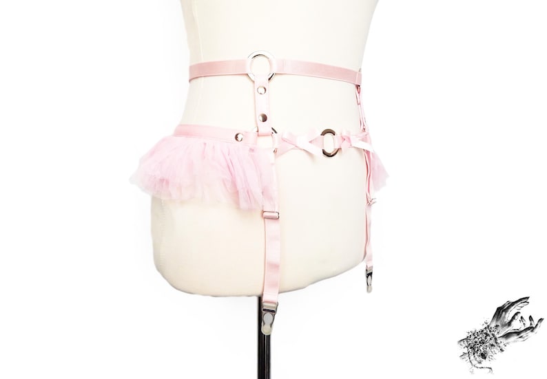 Adjustable pink vegan leather garter belt crafted for both functionality and style, catering to alternative fashion enthusiasts and BDSM communities alike. Featuring a central silver O ring, pink satin bow details, ruffled tulle accents at the side of the hips and pink elastic suspender straps, it’s a versatile garter belt that complements pastel goth and Japanese fashion, burlesque costumes or festival outfits. Displayed on a white mannequin bust to show the flattering, body conforming fit.