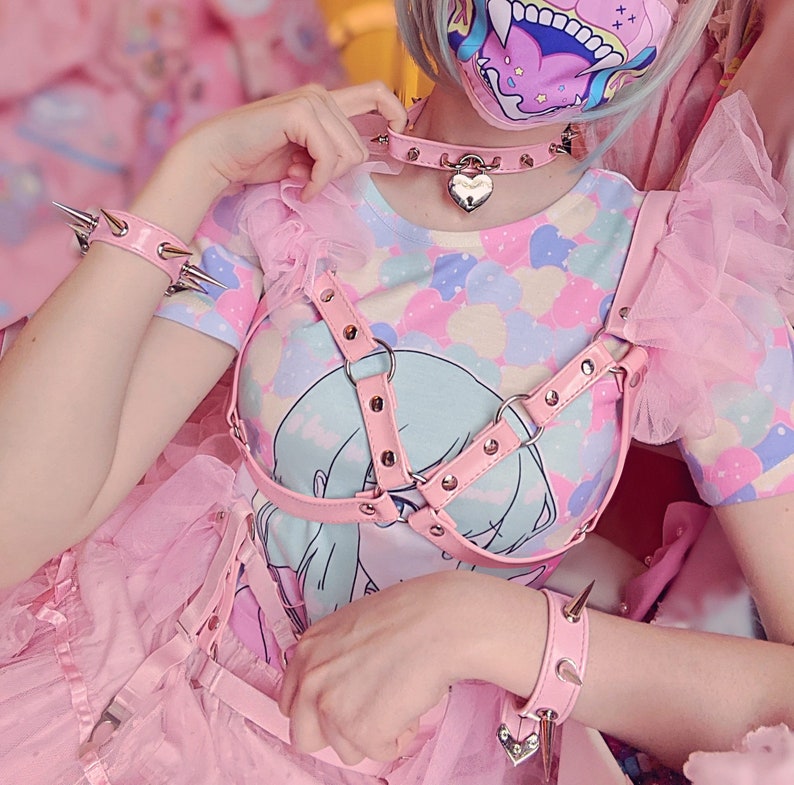 Modelled photo of the pink ruffled harness bra, styled with a matching pink garter belt, studded heart padlock choker, pastel blue and pink graphic t-shirt and pink tutu skirt for a Harajuku fashion aesthetic. The adjustable shoulder and bust straps at the back ensures a comfortable, versatile fit for all body types and sizes. Perfect for alternative fashion styles including pastel goth, kawaii and e-girl style, as well as BDSM inspired outfits, burlesque costumes, festival fashion and ravewear.